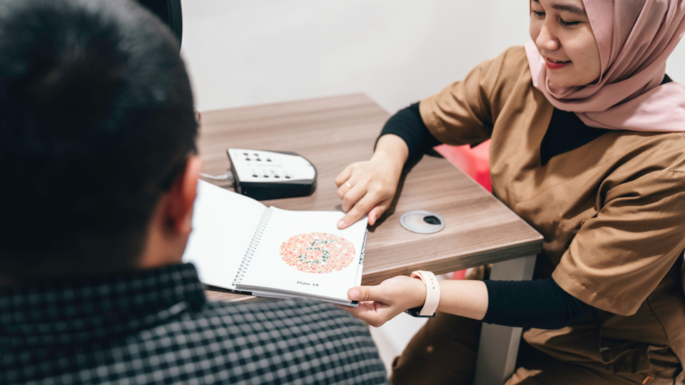 A female clinician presents a patient with an Ishihara colour vision test plate while seated at a desk