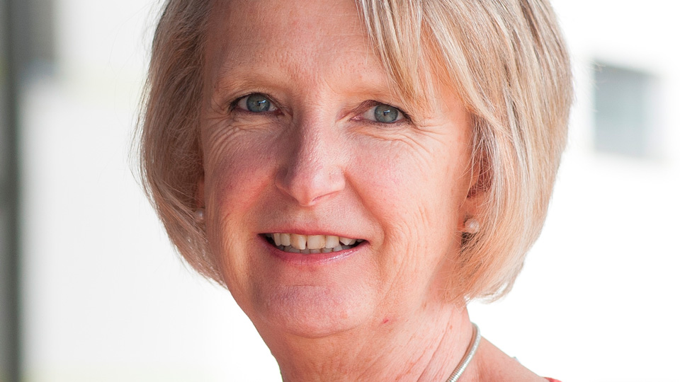 A woman with short, light grey-blonde hair smiles at the camera, wearing a red top and a silver necklace, photographed against a softly blurred, light background