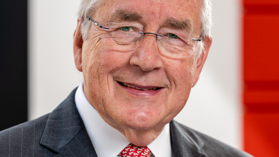 Head-and-shoulders portrait of an older man with short grey hair and glasses, smiling at the camera. He is wearing a dark pinstriped suit, white shirt, and a red patterned tie, with a softly blurred background featuring black, white, and red vertical sections