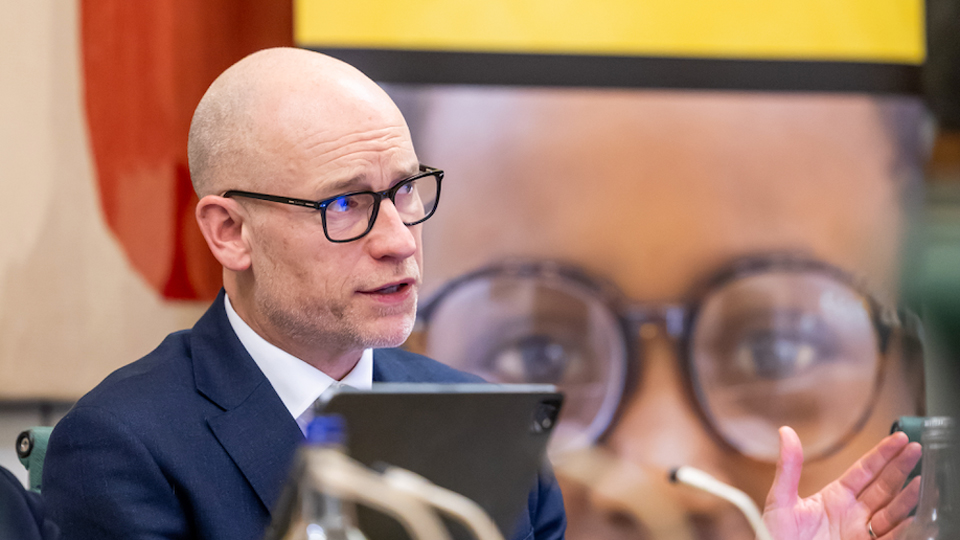 A man in navy blue suit and black glasses is talking on a panel. A banner in yellow and black behind him talks of the Value of Vision.