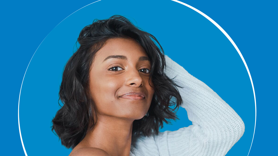 A young woman poses with her hand in her shoulder-length dark hair. A light blue-white knitted jumper slips off of one shoulder for an effect that is care-free. Behind her is a blue and white circle design