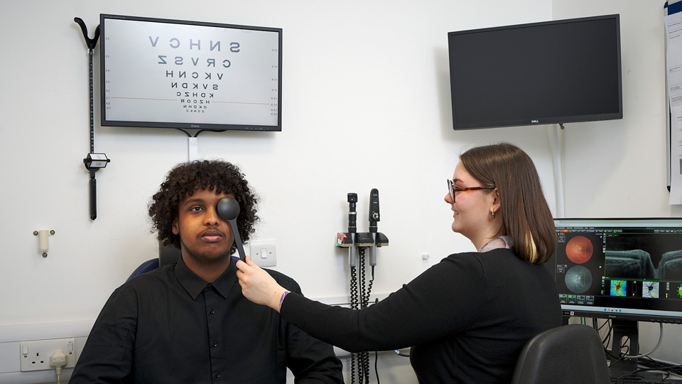A student practices a cover test on another patient in a clinical-styled environment. Above the patient is the letter chart