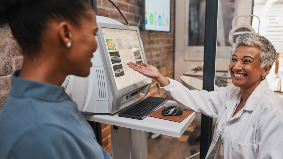 An eye care professional smiles to a patient as they gesture towards readings on a screen suggesting eye health indicators