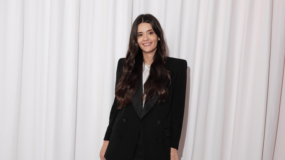 Patricia poses in an all white room wearing a long tailored black jacket with structured shoulders