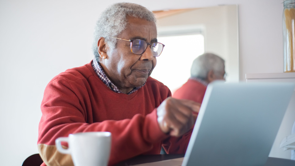 An older man is sat at a table using a laptop. He has a cup of coffee beside him and is using his finger to follow something on the screen. He wears glasses
