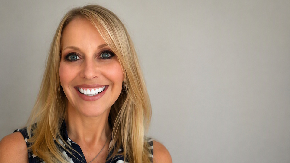 Head-and-shoulders portrait of a smiling person with long blonde hair and blue eyes, wearing a sleeveless navy and white patterned top, against a plain light grey background with copy space to the right