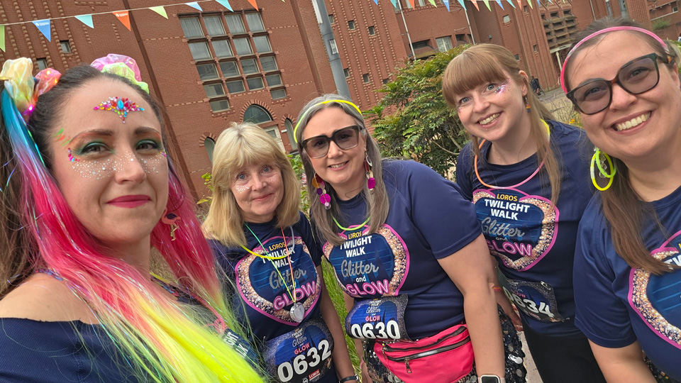 Five participants pose for a selfie before the LOROS Twilight Walk: Glitter and Glow event, smiling in matching navy event T-shirts with glow necklaces and glitter makeup. One person wears a bright rainbow wig and colourful face gems, while others accessorise with sequinned skirts, neon headbands and waist bags. The group stands outdoors in front of a large red-brick building decorated with bunting