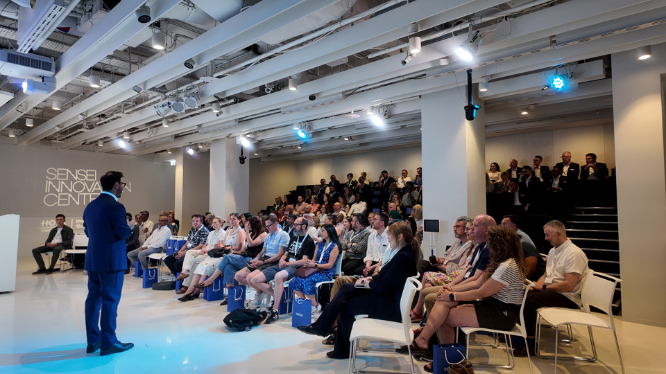 Jonathan Cohen in a navy suit stands at the front of a room of tiered seating welcoming attendees to the event. A projected screen in the background states Sensei Innovation Centre