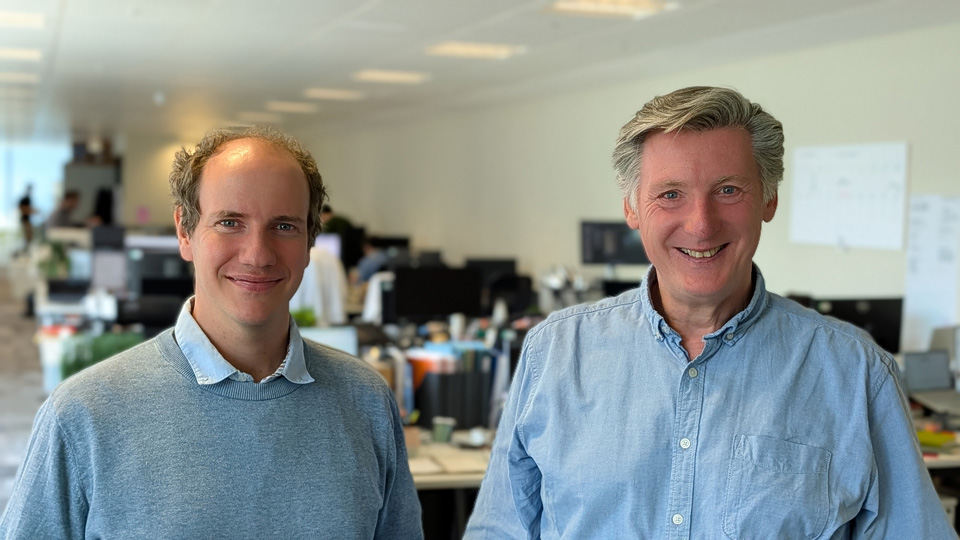 Dr Alistair Bounds in a blue shirt collar and grey jumper stands with Dr Dan Daly, who wears a blue linen shirt, in an office