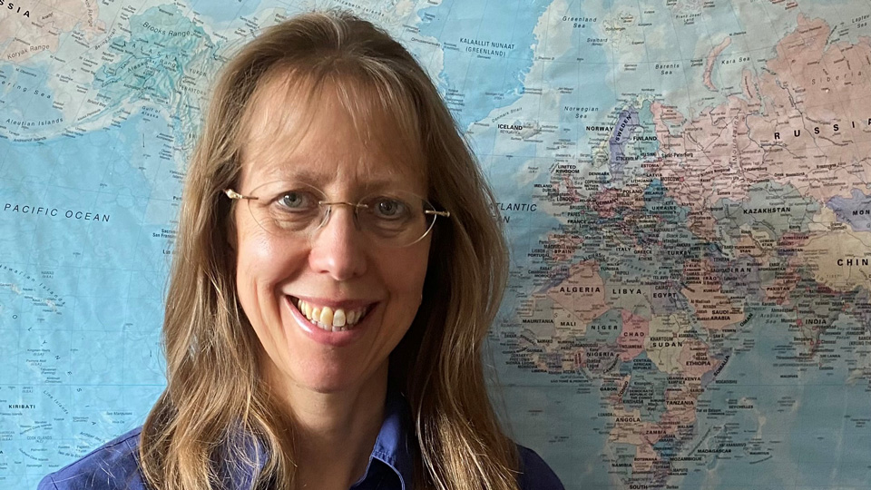 Anne stands in front of a world map, smiling for the camera. She has blonde-brown hair past her shoulders and wears a navy shirt