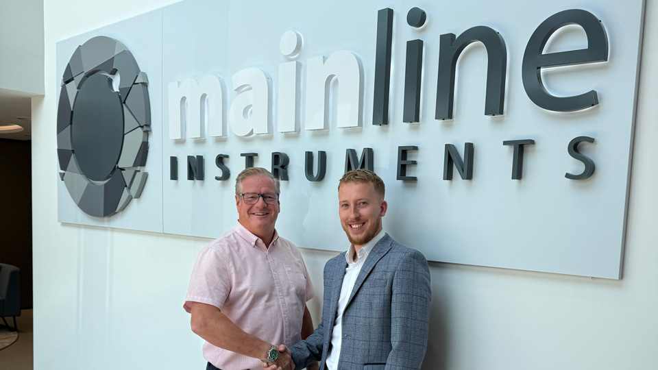 Graham Trevor and Jack Hawkins shake hands under a sign for Mainline Instruments. Graham wears a short-sleeved pink shirt and dark blue trousers, while Jack wears dark wash jeans, a white shirt and grey check blazer