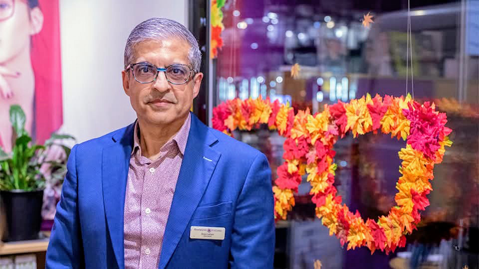 Deven wears a suit and stands by a floral arrangement in the shape of spectacles