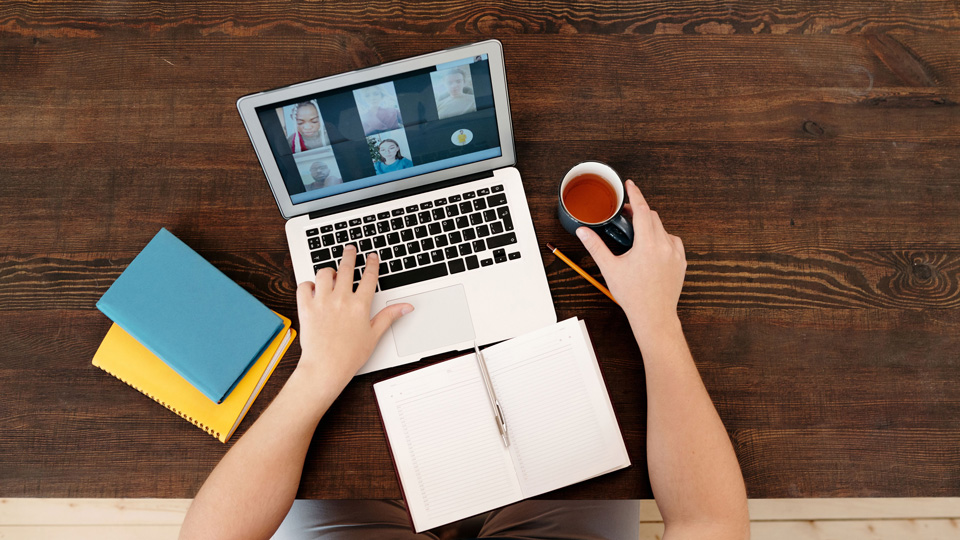A birds-eye-view of a man attending a virtual meeting, with notebooks in front of him. He types with one hand and holds a cup of tea in the other