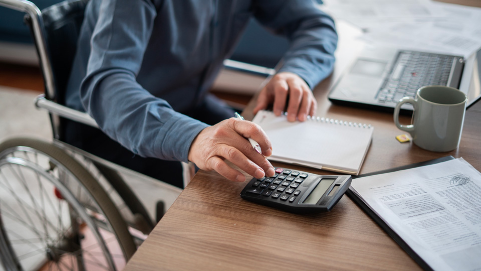 A man in a wheelchair uses a calculator and laptop while seated at a desk