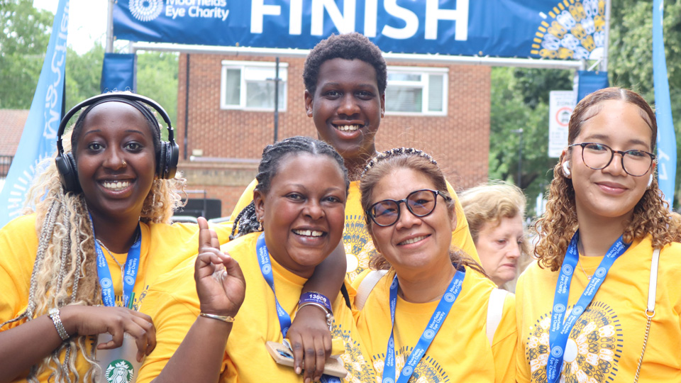Five participants wearing matching yellow event T-shirts and blue finisher medals smile for a group photo beneath a "FINISH" banner at the Moorfields Eye Charity event. One participant has an arm around another's shoulders, while others wear headphones or glasses. Trees, event flags and spectators are visible in the background, creating a celebratory finish-line atmosphere