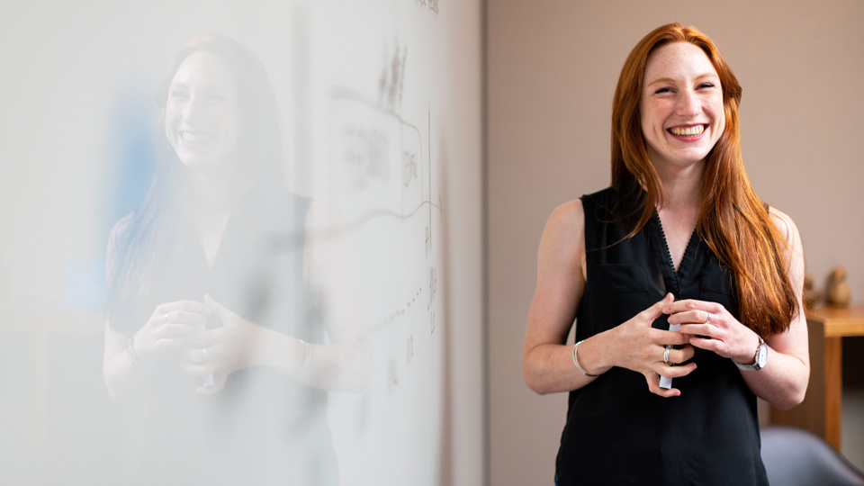 A redhaired woman in professional attire holds a pen and stands, smiling, beside a whiteboard