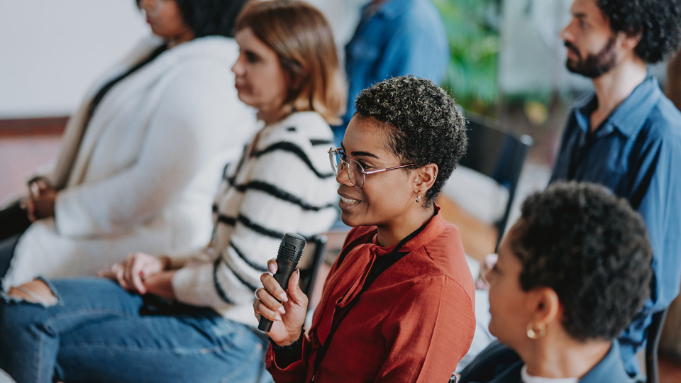 People sitting in a lecture with focus on a lady wearing glasses holding a microphone