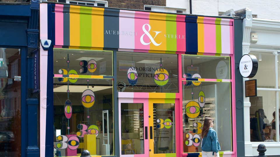 The shopfront of Auerbach & Steele on Kings Road High Street in Chelsea. To celebrate the anniversary of the practice, it has been painted in vibrant lines of mustard, lime, navy, and pinks. In the windows stained-glass style eyes and art-deco ‘30’ signs are arranged