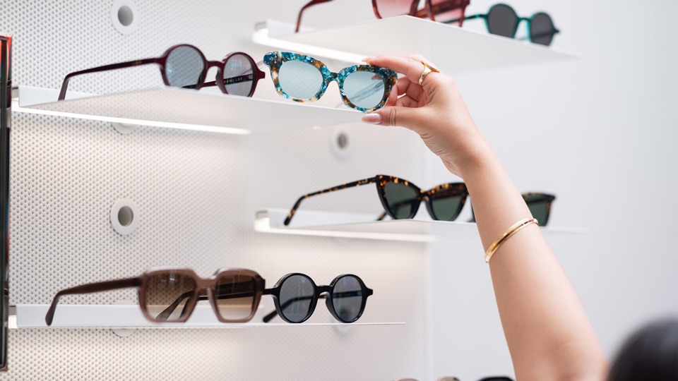 A hand reaches for a mottled blue-brown cat’s eye frame, on display in the practice. On white shelves around it are other stylish sunglasses