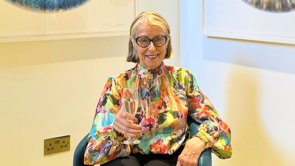 Gail is seated in a corner of practice holding a glass of champagne. She wears a long sleeved blouse in a vibrant watercolour and rounded cat’s eye style glasses. On the two walls behind her are large canvases of iris art
