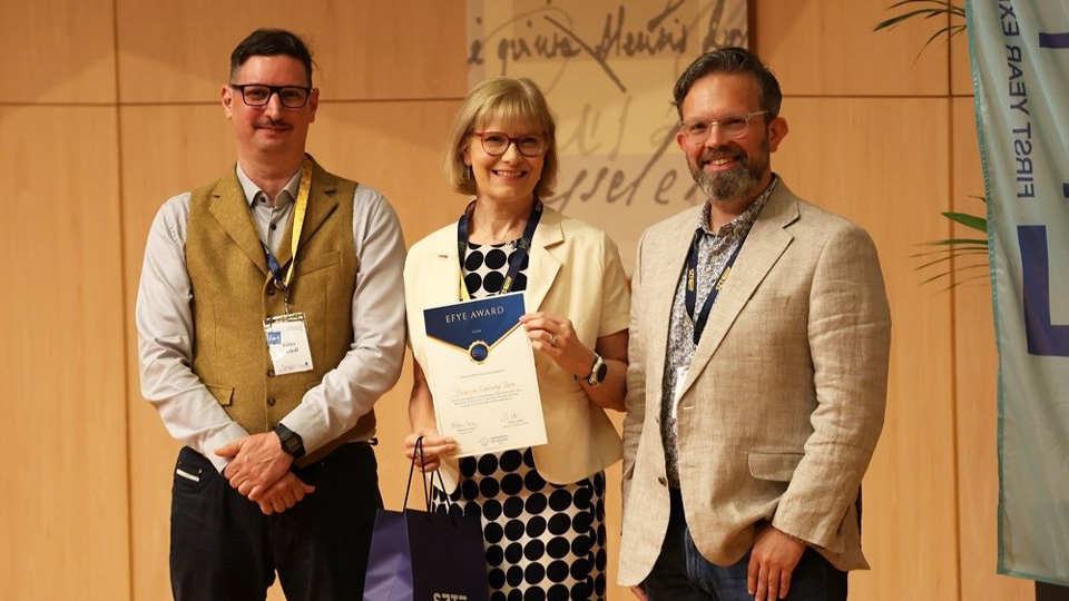 Three people stand on stage in formal attire. In the centre, Catherine holds a certificate for the EFYE Team award