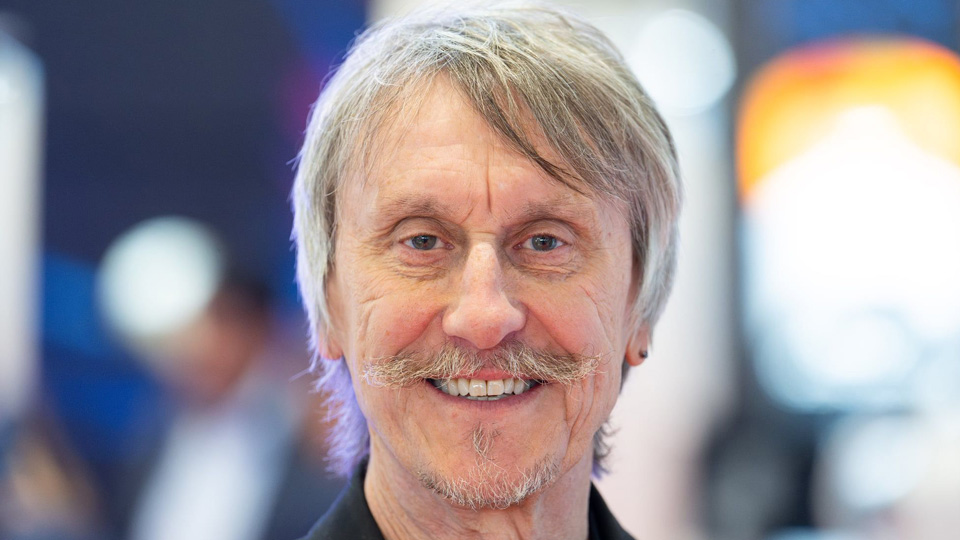 A close-up headshot of Brian in a black t-shirt and blazer. His gray hair is cut long and he has a moutsache and goatee