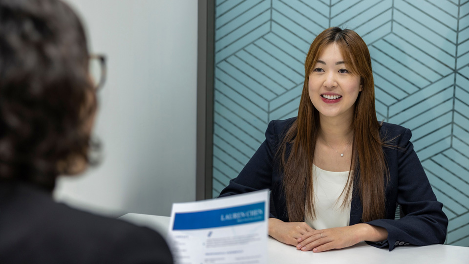 A young woman in a smart blazer is seated on the far side of a circle table, she appears engaged and enthusiastic. In the foreground, blurred, a person holds a document that appears to be a CV