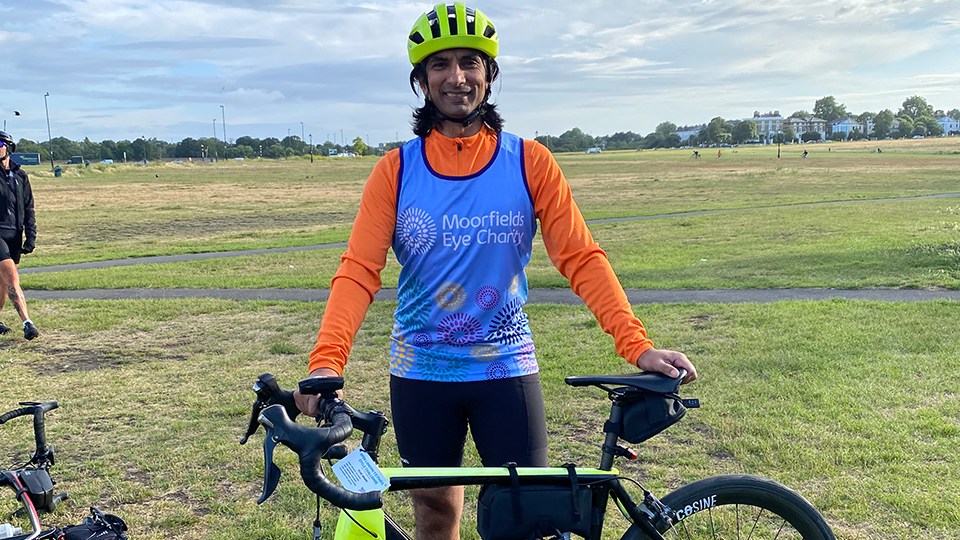 Vijay wears a Moorfields Eye Charity vest over an orange cycling jersey and a yellow helmet. He stands behind his bike, which is fitted with yellow reflectors. The background is an open grassy park