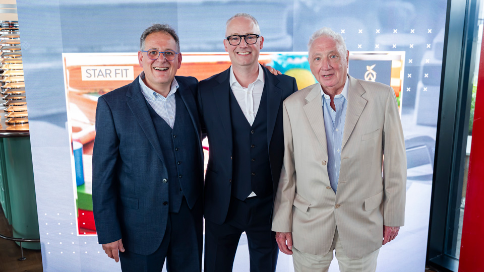 Three men stand together smiling in celebration, all wearing smart suits. They stand in front of a light board with an advertisement for an eyewear product. <Image title> n/a