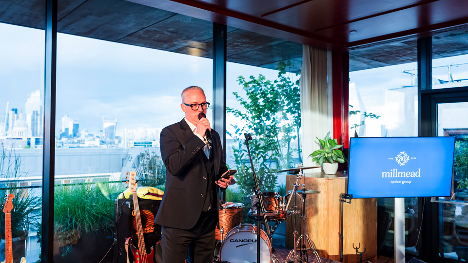 James Conway is speaking into a microphone. He wears a black suit. Behind him is a screen with the millmead logo, instruments, and the London skyline through large windows