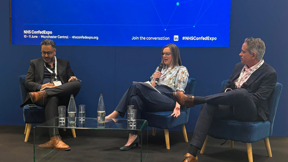 Speakers sit on a panel in front of a blue screen at NHS Confed Expo