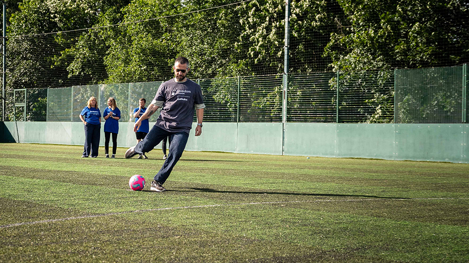 On a football pitch, a man wearing a grey Freedom Vision t-shirt and darkened goggles lifts his foot back to take a penalty kick
