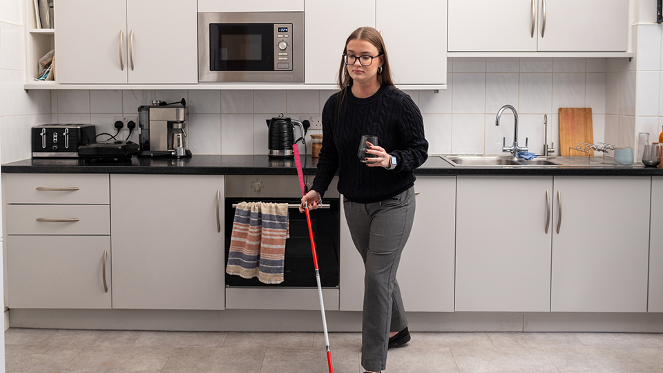 A full shot of a young woman with dual sensory loss, walking away from a kitchen holding a glass of water in a clean, bright kitchen. She is holding her red and white cane, providing essential mobility, safety, and independence