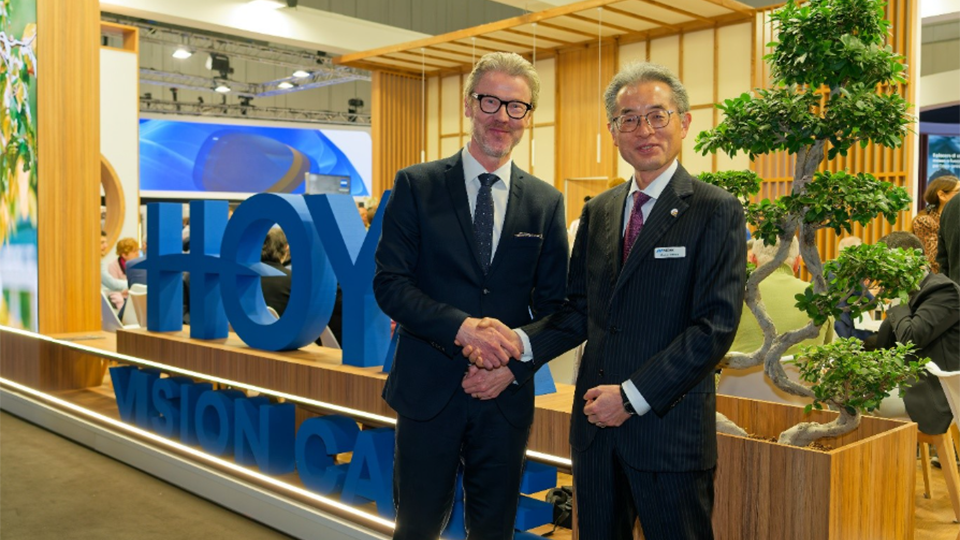 Two men dressed in black suits shake hands in front of a sign for Hoya Vision Care