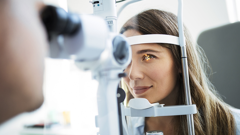 A young woman receives an eye test, her face positioned in a slit lamp device for assessment with a light shining into one eye