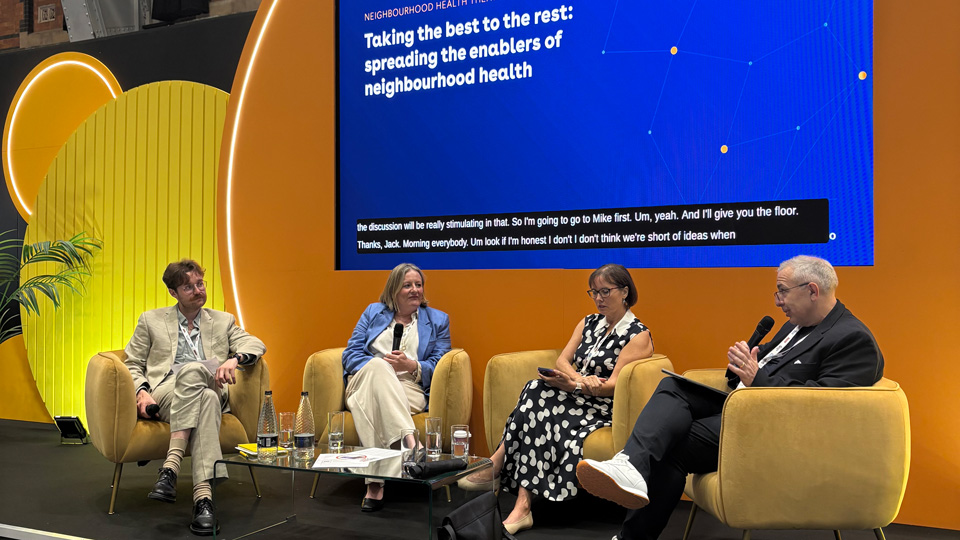 Four speakers sit in mustard-yellow armchairs on a conference stage at ConfedExpo, taking part in a panel discussion. Behind them, a large blue NHS-branded screen displays the session title, “Taking the best to the rest: spreading the enablers of neighbourhood health.” The stage features bold circular yellow-orange set pieces and is located in a spacious hall with exposed brick walls, tall windows and steel beams. A glass coffee table with water bottles and papers sits between the panelists