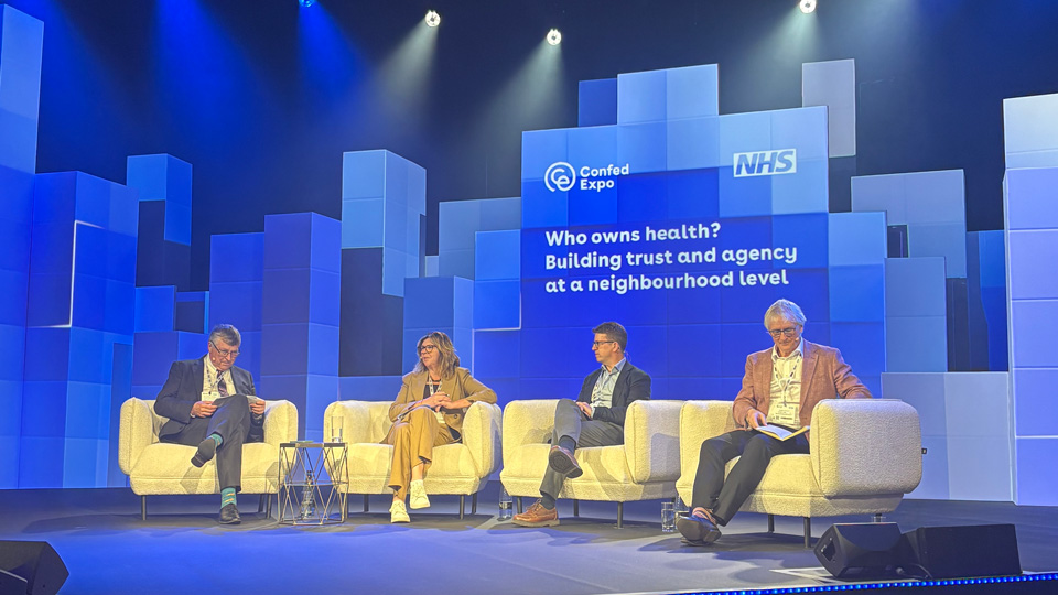 Four panelists sit on cream-coloured sofas on a conference stage at ConfedExpo, beneath blue and white lighting. A large NHS-branded screen behind them displays the session title, “Who owns health? Building trust and agency at a neighbourhood level.” The backdrop is made up of stacked geometric panels in varying shades of blue and white, creating a cityscape-like effect. Small tables with water glasses sit between the speakers as they take part in the discussion