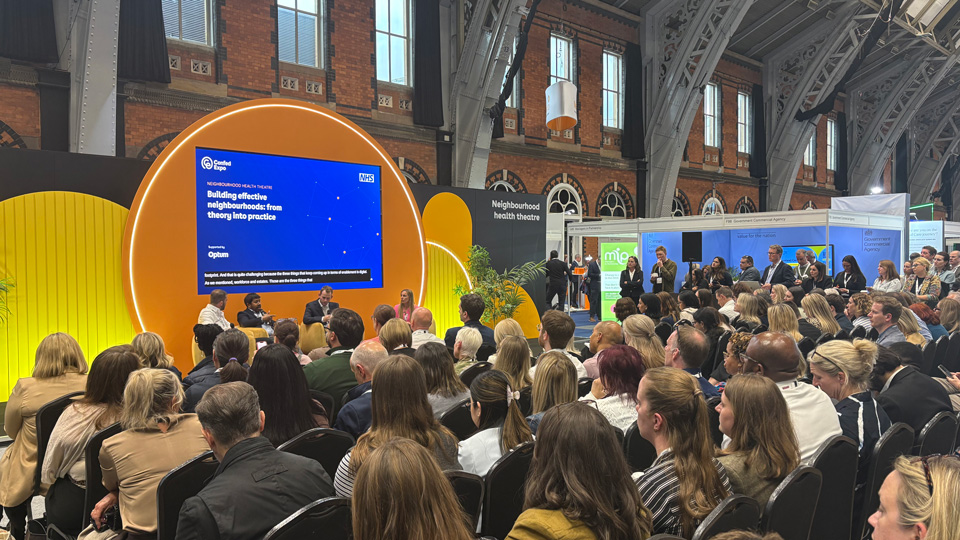 A crowd of seated people look towards a stage where a panel is talking in front of a screen