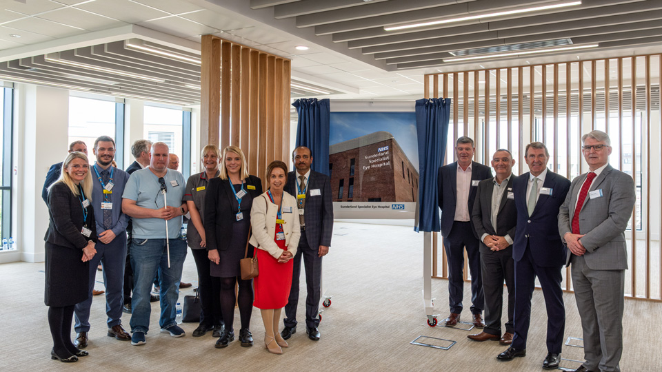 A large group gather around a sign displaying the new Sunderland Specialist Eye Hospital, on either side of the display are blue curtains which have been pulled back in a reveal.