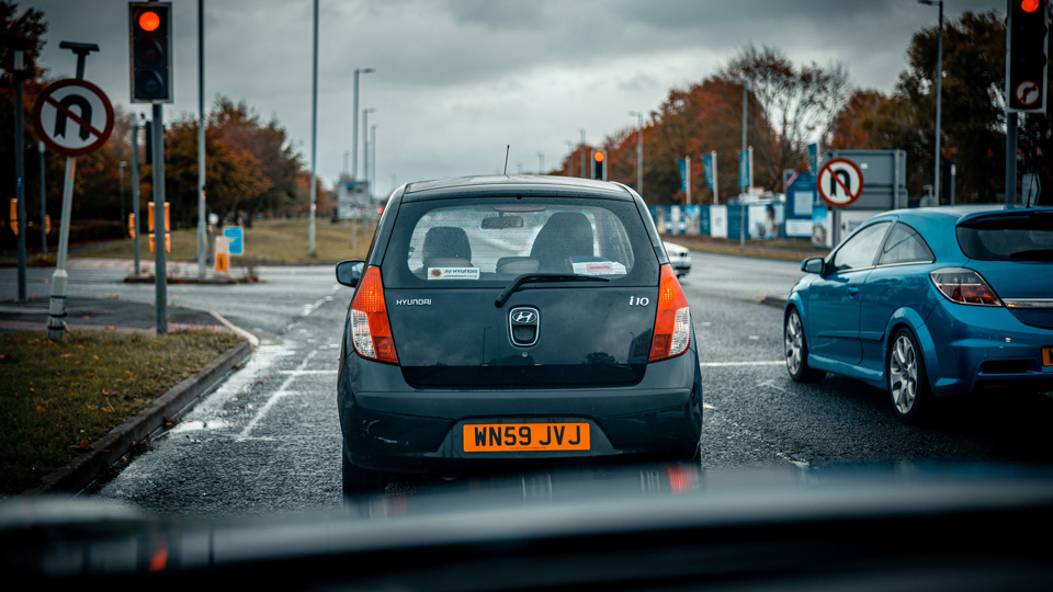 Taken from inside of a car, the windscreen wipers just visible at the bottom of the frame, the photo captures traffic waiting at lights