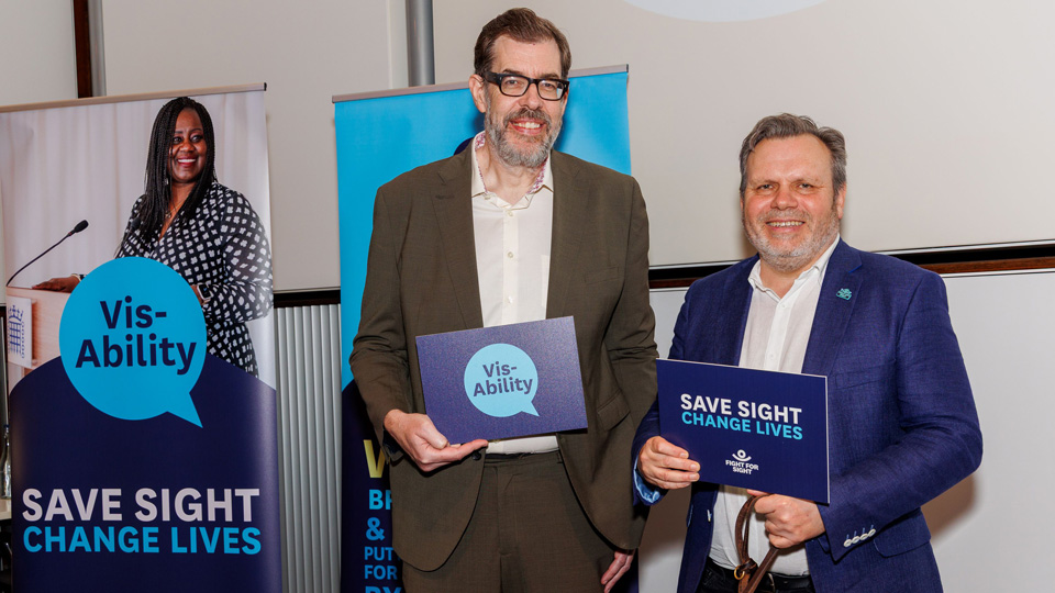 Richard Osman and Keith Valentine pose with signs for Vis-Ability and Change sight change lives messages in front of FIght for Sight banners
