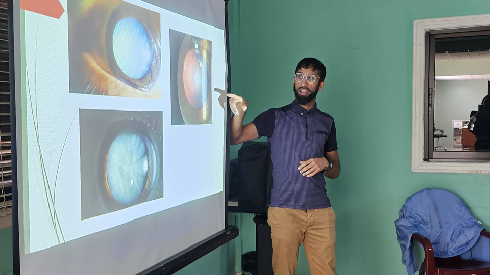 Benji stands at the front of a teal painted room. He is wearing brown chino trousers, a navy polo shirt and trainers. He points at an overhead projector screen which displays images of cataracts