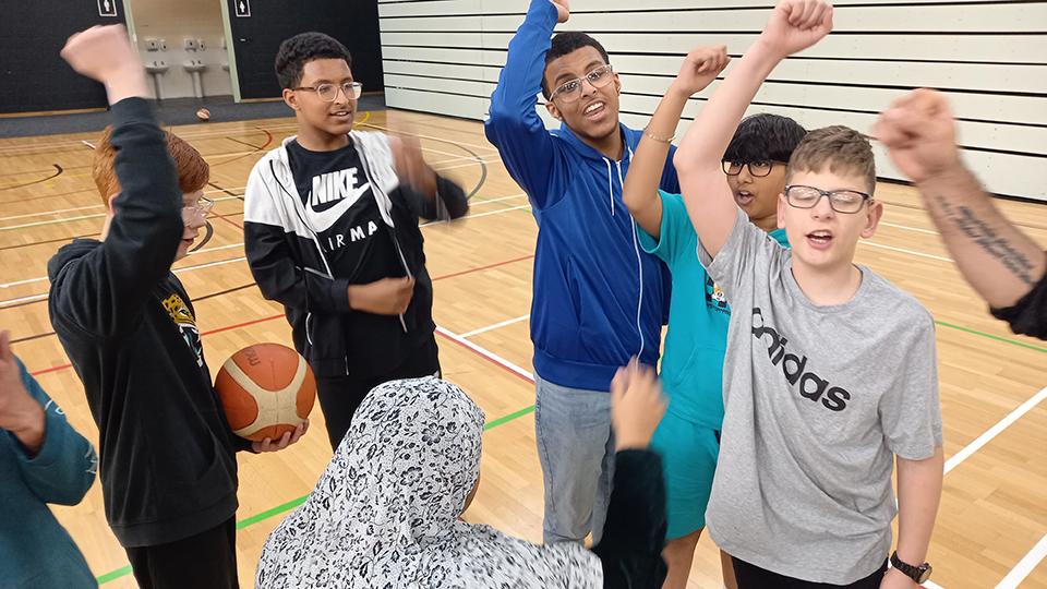 A group of young people stand in a circle on an indoor sports court, raising their arms together in a team cheer. One boy holds a basketball while others smile and shout with their hands in the air. The group appears energetic and supportive, gathered on a wooden gym floor with court markings and sports hall walls in the background