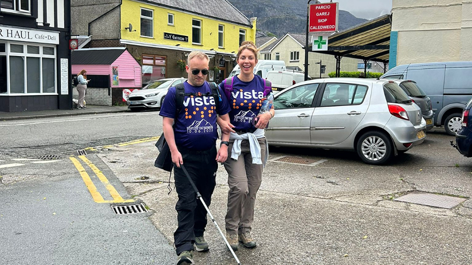 A man using a white mobility cane walks along a village street with a woman beside him, both wearing purple Vista charity T-shirts. They appear to be taking part in a walking challenge, with mountains rising in the misty background and small shops and parked cars lining the road. The woman smiles toward the camera while the man focuses ahead as they walk together