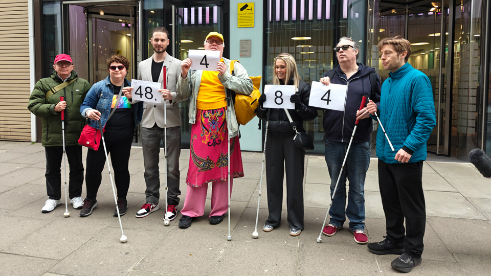 Image shows from left to right, cane users Geoff Smith, Charmaine Ashpole, Rob Sears, Frankie Brady, Claire Sisk, Mike Wordingham and Tom Skelton standing in front of the Department of Health and Social Care building in Westminster holding up large pieces of paper with 48, 4, 8 and 4 on them. The numbers represent the number of signatures gained for RNIB’s Vision Rehabilitation petition.