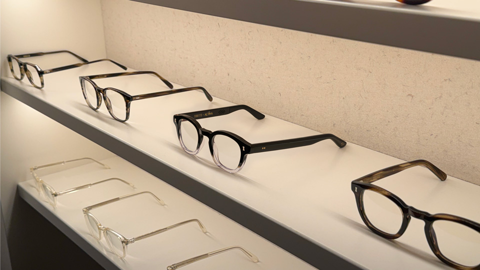 A shelf of Cubitts eyewear with the Cubitts x Altina frame in the centre. The frame is black with an ombre effect making the bottom of the frame front clear. One of the inside of the arms is visible, revealing a pattern indicating a speaker
