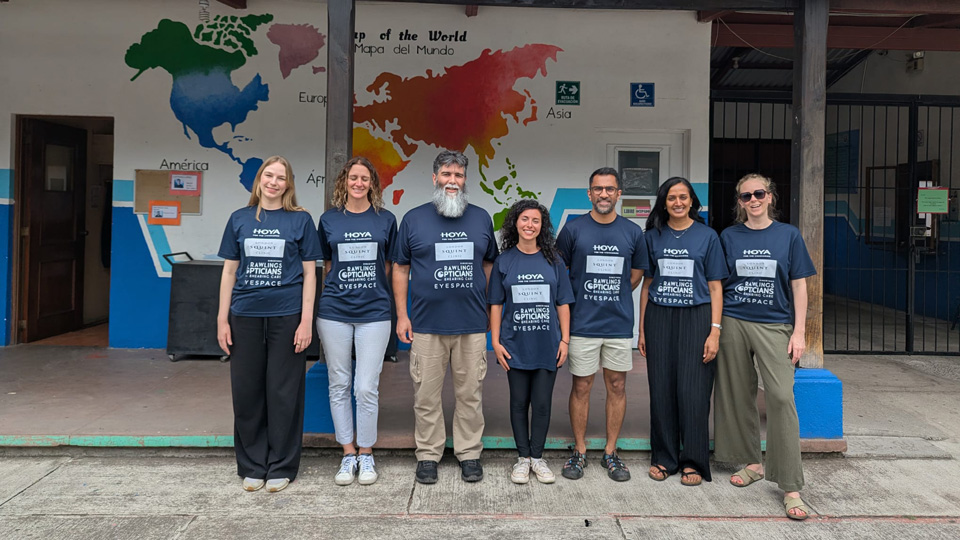 Seven people stand together outside of a building with a large watercolour style world map painted on the side. The team are wearing t-shirts branded with Hoya, London Squint Clinic, Rawlings Opticians & Hearing Care, and Eyespace