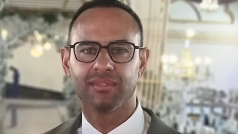 Naheem wears a crisp white shirt and a green suit jacket, and spectacles with a bold black rim