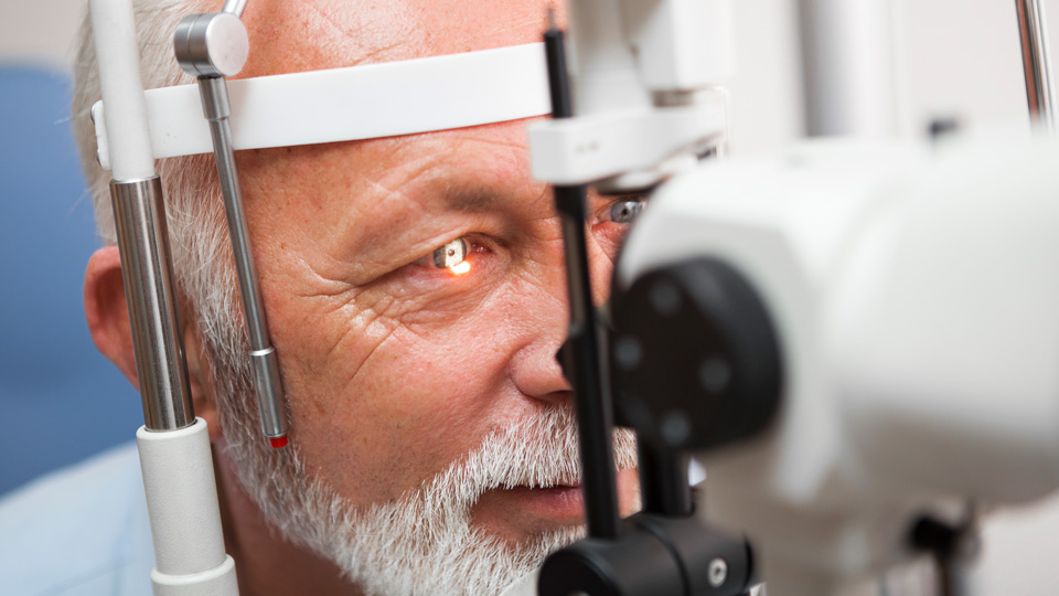 A close up of an older man receiving an eye test. His face is positioned on the chin rest and a light shines in his eye