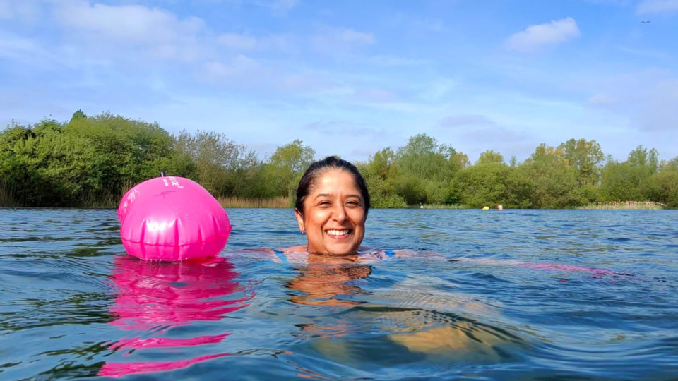Kavita is swimming in the blue waters of a local lake. She grins joyfully at the camera. The sky is blue and there are trees in the background.